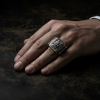 A close-up of a hand wearing a large square signet ring with an engraved crest on a dark textured surface.