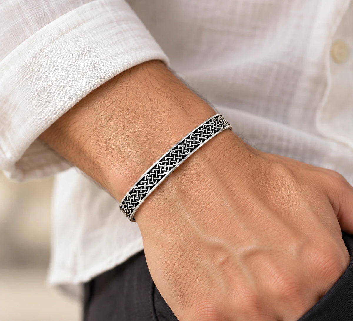 A close-up of a man's wrist wearing a silver-toned metal cuff bracelet with an intricate black woven geometric pattern, styled with a white linen shirt.