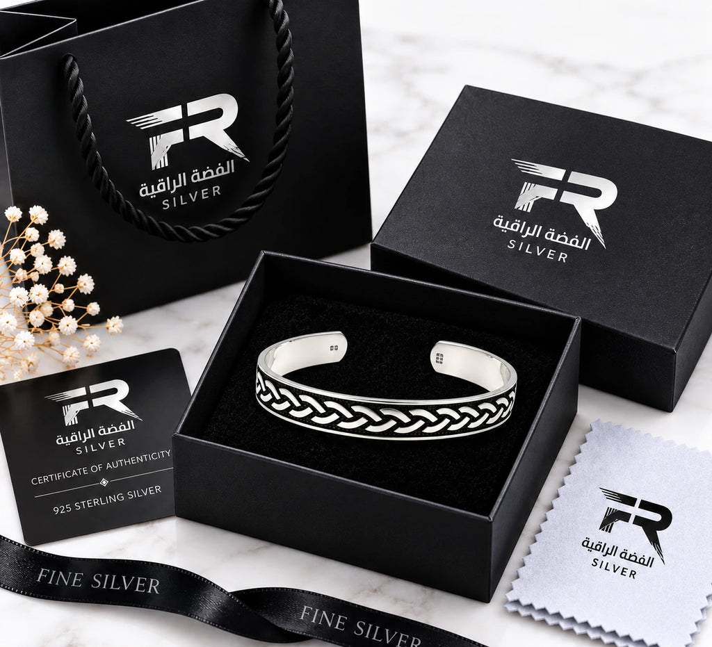 A sterling silver cuff bracelet with an engraved braided pattern, presented in an open black gift box alongside its packaging, including a black shopping bag, certificate of authenticity, and microfiber cloth, all branded with the 'Raffaella' logo.