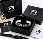A sterling silver cuff bracelet with an engraved braided pattern, presented in an open black gift box alongside its packaging, including a black shopping bag, certificate of authenticity, and microfiber cloth, all branded with the 'Raffaella' logo.