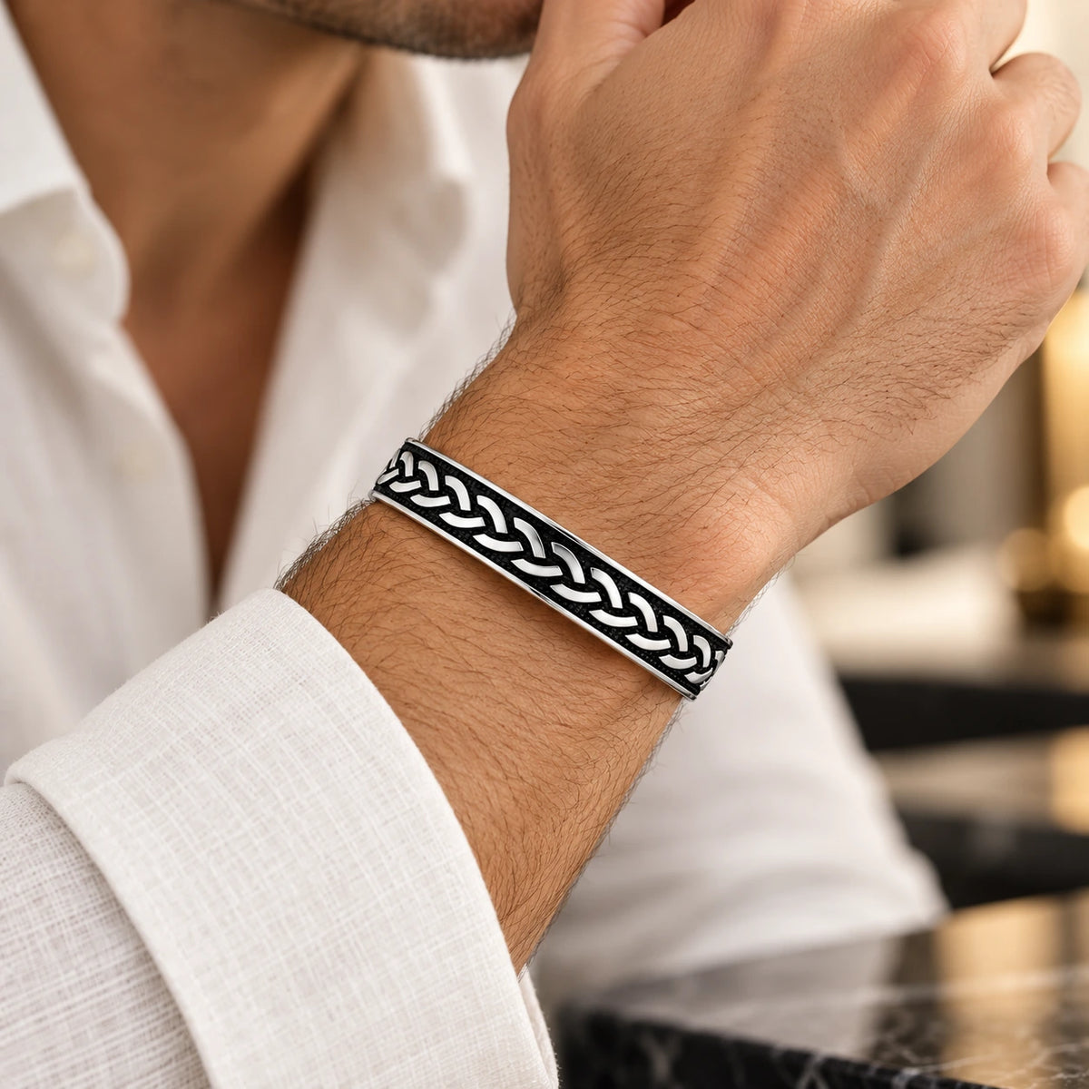 A close-up of a man's wrist wearing a silver-toned metal cuff bracelet with a black and white braided pattern, paired with a white linen shirt.